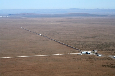 Aerial photo of LIGO facility. White buildings are visible in the lower-right corner, and two long pipe-like structures extend a right angles from the buildings across an open, flat landscape. Mountains are in the distant background.