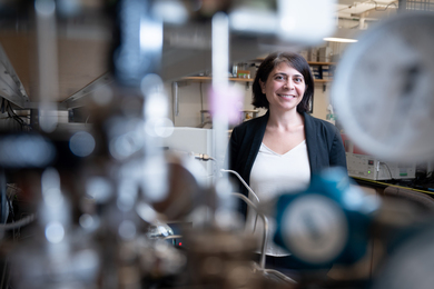 Desirée Plata smiles in the lab and is seen through blurry metallic equipment in the foreground.
