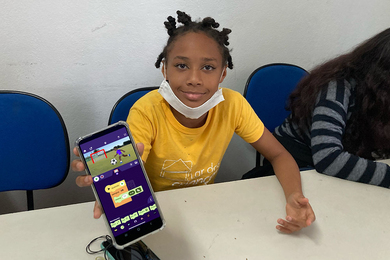 A child seated at a table in a white room holds a smartphone up to the camera to show off their soccer project.
