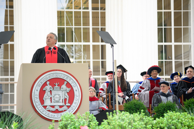 Noubar Afeyan speaks at a podium with the MIT seal on the front. Faculty and administrators in academic regalia are seated next to him.