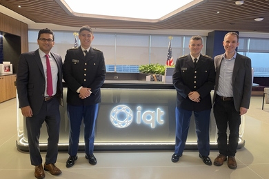 Aneal Krishnan, William Cruz, Alexander Edwards, and David LoBosco pose in front of a desk with a backlit “IQT” logo. Cruz and Edwards wear military cadet uniforms.