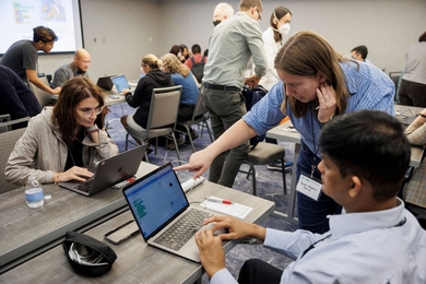 More than a dozen people sit around shared tables with laptops running App Inventor