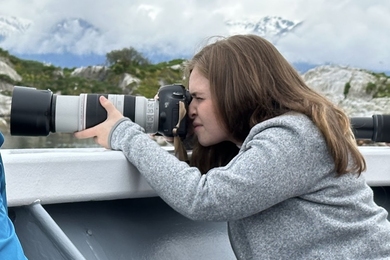 Side view of Sophie Hartley squinting into the viewfinder of a camera with a very large telephoto lens, with cloud-shrouded mountains in the background..