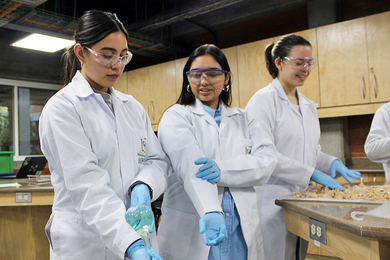 Lourdes Figueroa and two students, all in lab coats, stand by a bench, practicing how to hold a glass container