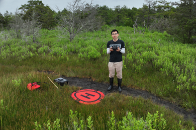 Ernie Lee stands in a dense marsh holding a controller as a drone takes a photo of him. 