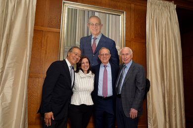 Behind the four people, the large portrait shows L. Rafael Reif in a blue seat smiling, with MIT columns in background.