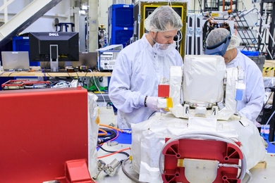 Two individuals in cleanroom attire examine an optical communications system payload. 