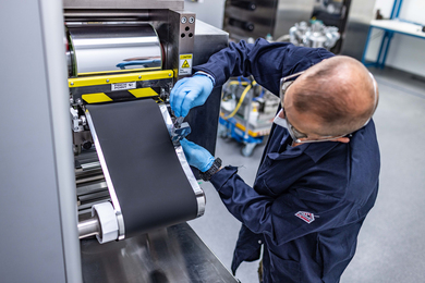 A man works on a large machine that uses rolls of silver and black material.