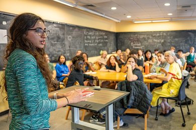 Angelina (Angie) Ayoubi stands at a lectern in a classroom with about 20 other students listening to her