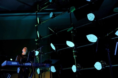 On a dark stage, Jordan Rudess performs on keyboard in front of a kinetic sculpture featuring a grid of white petals