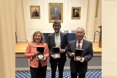 Daniela Rus, Takeo Kanade, and Vijay Kumar pose together displaying medallions held in boxes. Behind them are portraits of Ben Franklin and other American founders.