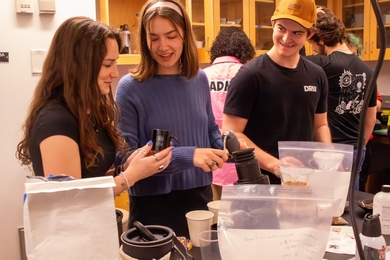A student holds an espresso cup at a lab table while talking to her two classmates.