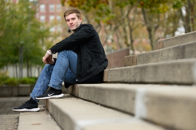 Taylor Hampson, wearing jeans and a light jacket, sitting outdoors on concrete steps with trees and a brick building in the background