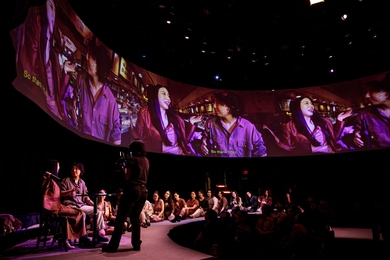 Two actors sit at a small round table, their images projected multiple times on a large circular screen above them. Some 20 others sit on the floor in a semicircle behind them.