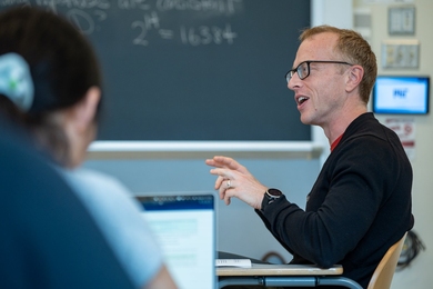 Brian Hedden sits at a desk in a classroom as he speaks to students seated around him.