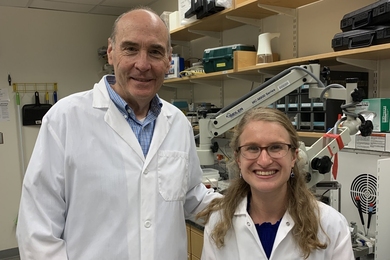 Mark Bear and Sara Kornfeld-Sylla in white lab coats pose in front of a laboratory bench