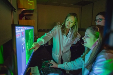 Professor Prescott, wearing a white lab coat, points to a microscope image on a screen. Two watching students are bathed in green light from the monitor