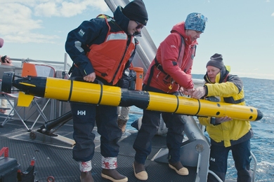 Ella Wawrzynek, Madeline Miller, and David Whelihan in winter clothing on a ship prepare to lower a long underwater vehicle into the ocean.