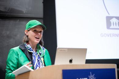 Georgina Campbell Flatter, wearing a bright green jacket and matching baseball cap, speaks at a lectern.