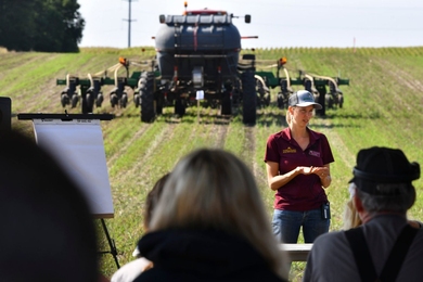 Nora Hertel stands in front of a tractor in a field, speaking to listeners in the foreground.
