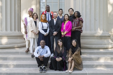 12 people, some with traditional dress from various countries, pose on the steps of an MIT building with columns behind them