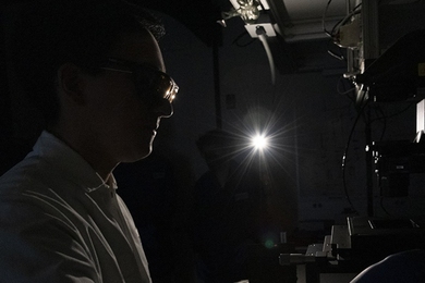Silhouetted close-up profile of a lab member, lit from behind, glancing through a mircoscope in a darkened room.