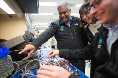 Anantha Chandrakasan smiles and points to a computer chip, alongside two grinning graduate students