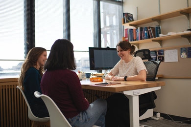 Anna-Christina Eilers sits at her desk, smiling at two female students who sit facing her