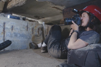 Niki Fandel, wearing a hard hat, sits, rather squished within a cramped concrete and dirt floor space, taking a photo with a large lens