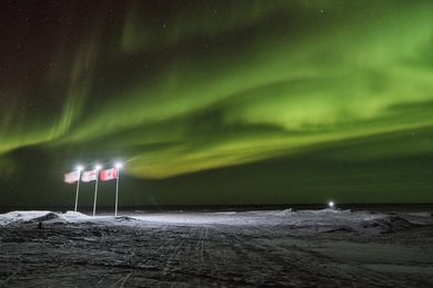 At night, 3 illuminated flags planted in Arctic ice wave against a backdrop of the northern lights