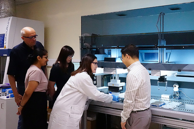 A woman in a lab coat operates equipment on a lab bench while four others look on