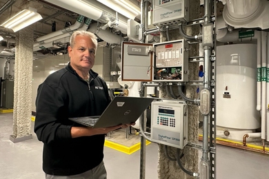 Fran Selvaggio holds an open laptop, standing beside equipment and pipes in a mechanical room.