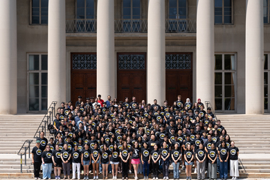 More than 100 BWSI students wear matching black shirts with the BWSI logo and stand on steps outside of an MIT building with columns in the background. 