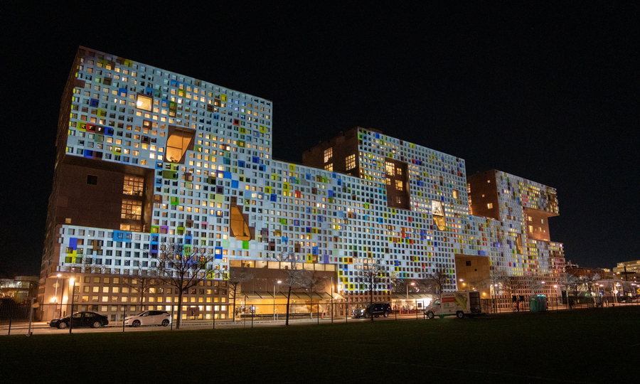 Photo of a multicolored art installation at night on the facade of Simmons Hall, a rectangular dorm with a brushed metallic gray facade, half a dozen large cutouts, and many small square windows