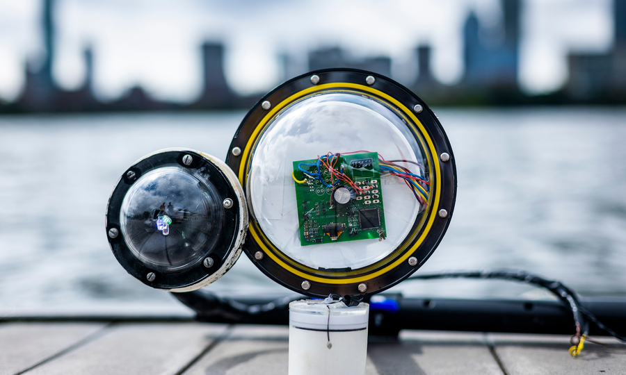 A prototype camera, with 2 bulbous lenses, a circuit board, wires and white cylinder, sits on a dock with the Charles River and Boston skyline in blurred background. 