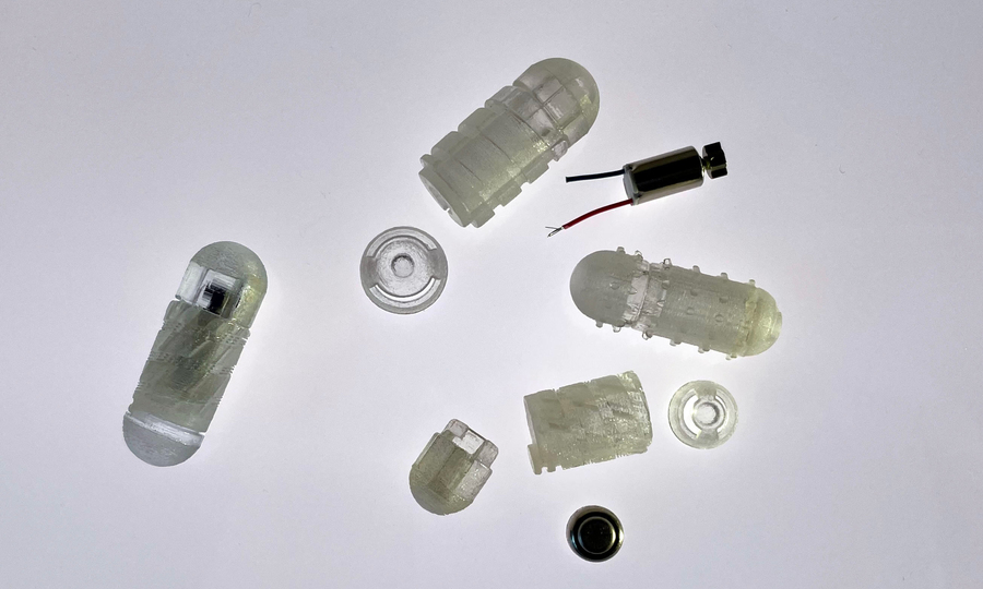 Nine pieces of the drug capsule prototype are on a white background. Some pieces are pill-shaped and made of translucent material. Some are threaded like a screw.