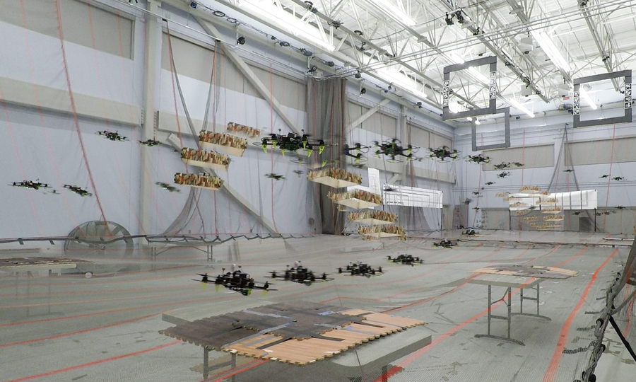 Combined video stills show the pathways of four flying drones in a large auditorium. The four drones avoid two flying obstacles, which are drones with a camouflage box around then.