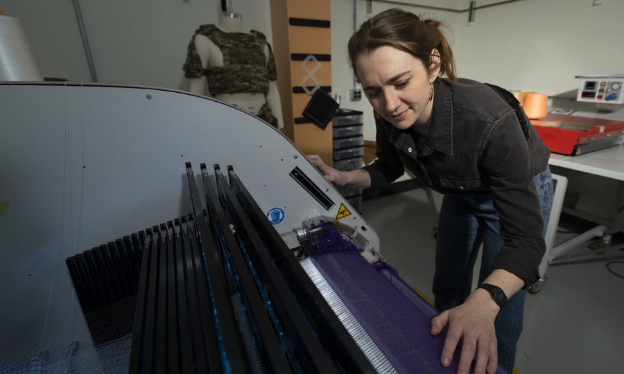 Erin Doran stands over a textile weaving machine demonstrating how a fabric is woven on it.