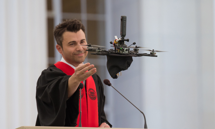 Mark Rober has attached his graduation cap to a flying drone, and he gestures to it as it flies away.