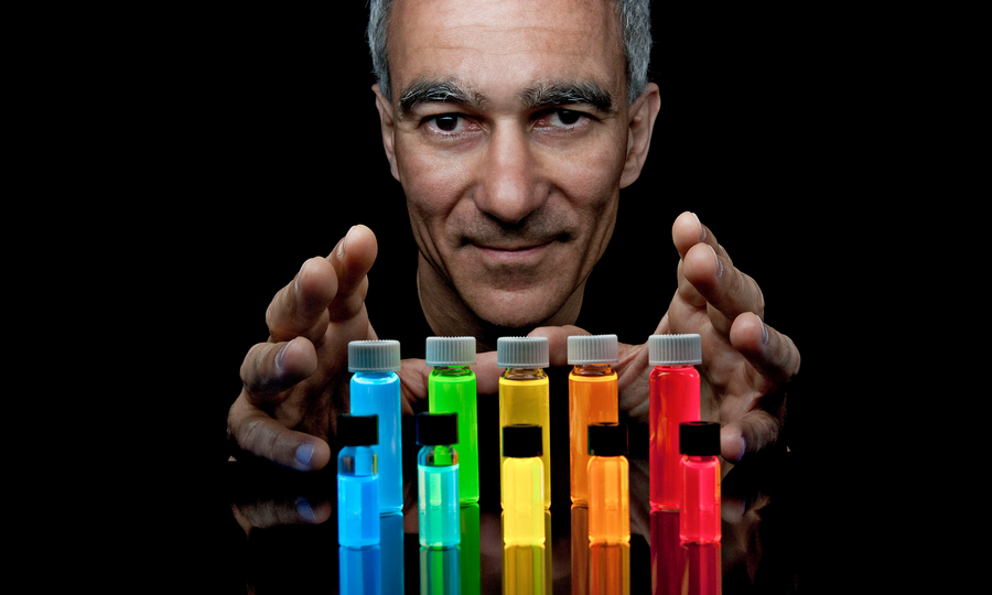 Moungi Bawendi's head and hands appear against a black background and over a rainbow-colored row of vials
