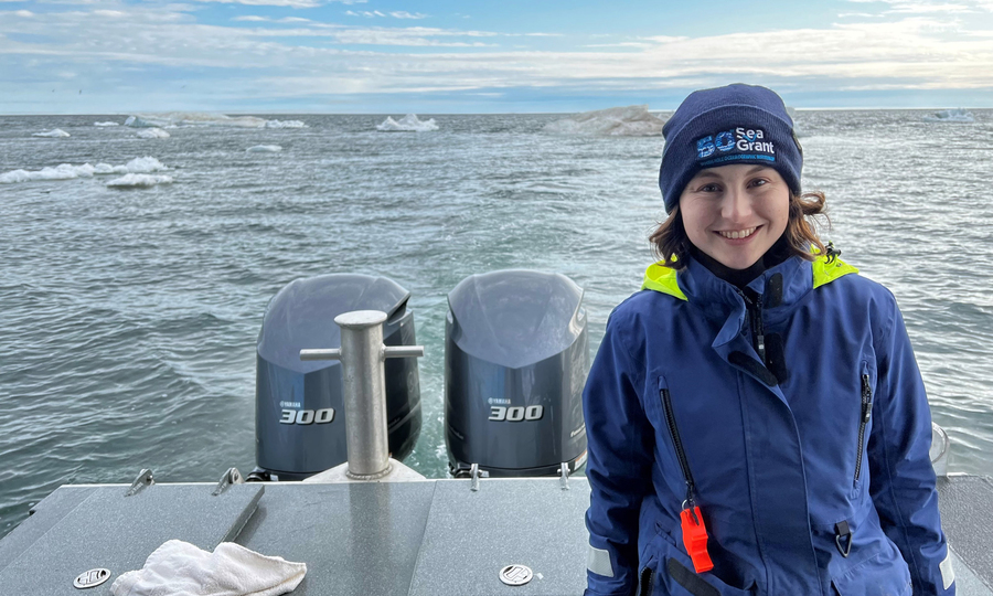 Emma Bullock smiles while near the back of a boat and wearing waterproof gear, with the ocean and sky in background.