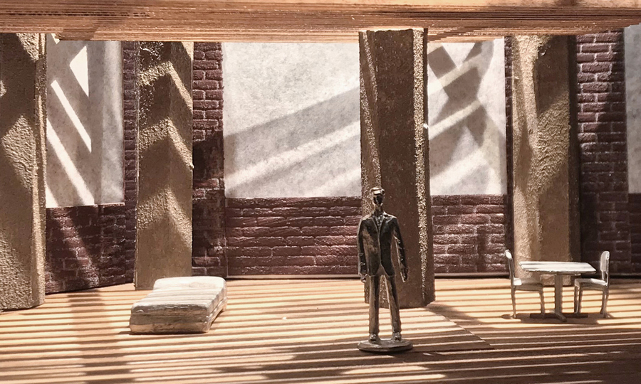 A small model shows a wooden man in a sparse room, with dramatic lighting from the windows.