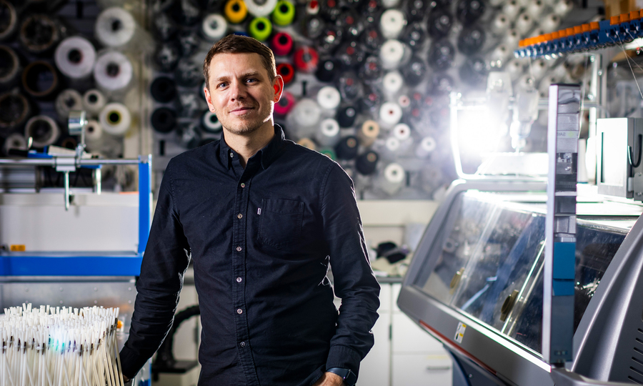 Skylar Tibbits stands in the lab with a wall of colorful spools behind him.