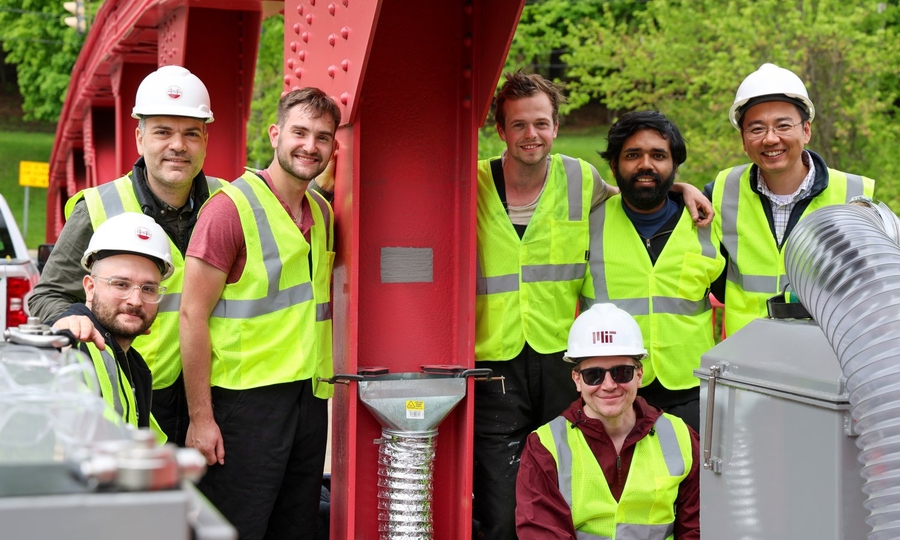 Seven men wearing safety yellow work vests and hardhats pose by a metal pillar that has a small gray patch in its center.