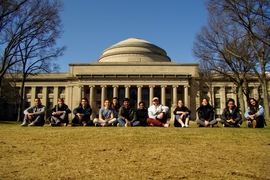 Twelve students sit cross-legged on a lawn in front of MIT's Great Dome during winter