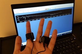 Photo of a hand with electrodes around two fingers, in front of a computer screen