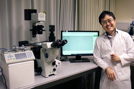 Photo of Kerwin Kwek in a labcoat, standing next to a computer, a microscope, and a 3D printer