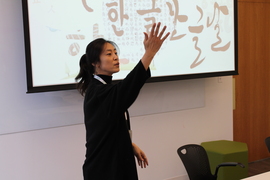 Photo of Hee-Jeong Jeong standing in a classroom, in front of a screen with Korean text, with her hand raised