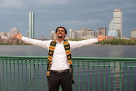 Photo of Mussie Demisse with arms outstretched, standing in front of the Charles River and Boston's skyline
