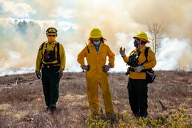 Photo of three people in yellow hard hats and jumpsuits standing in a field in front of smoke
